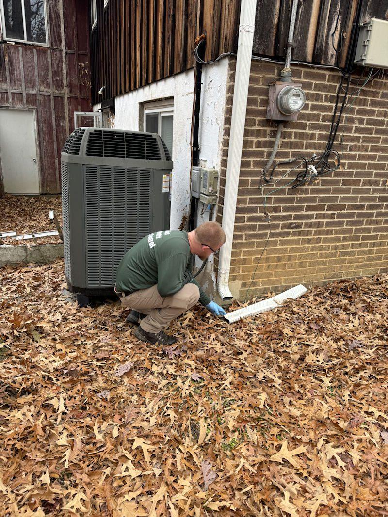 A Planet Friendly Pest Control technician checking exterior points around an Owings home — the HVAC unit, downspout and foundation line